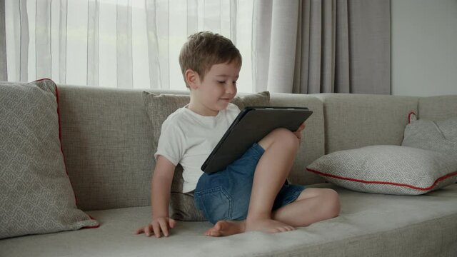 Cute preschool kid boy using digital tablet technology device online education sitting on couch at home. Children tech addiction concept. Child with gadget.