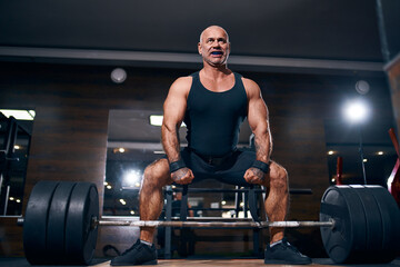 Fototapeta na wymiar Emotional Older sportsman preparing to exercise deadlift with barbell while on cross training in a gym.