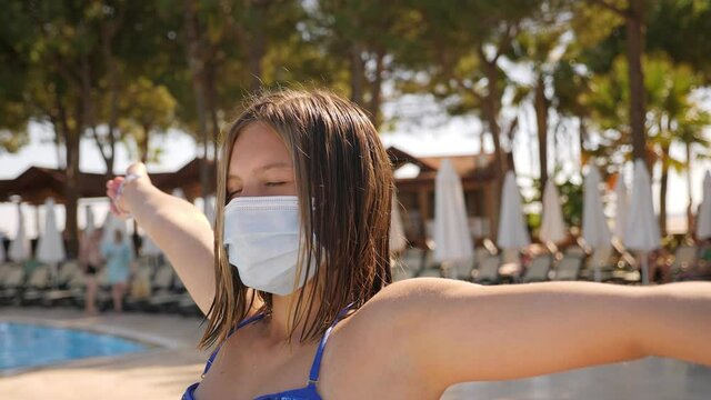 A teenage girl in a swimsuit and a medical mask is resting by the pool, she spreads her arms to the sides. Safe rest during coronavirus infection.