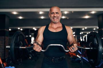Plakat Front view of older bodybuilder doing exercises with barbell at biceps in gym. Bodybuilding concept