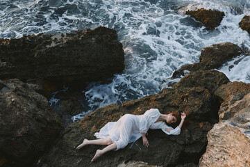 Barefoot woman in a secluded spot on a wild rocky coast in a white dress view from above
