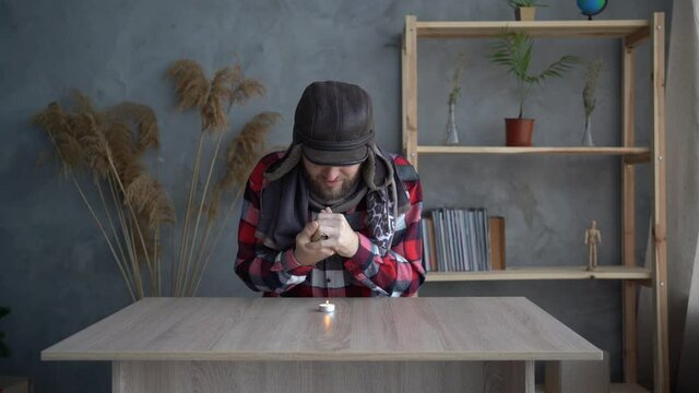 A Caucasian bearded man dressed in a warm winter hat and a scarf sits at home at a table and warms his hands from a burning candle. Concept of no heating in winter at home.