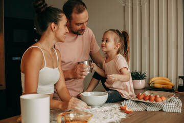 Obraz na płótnie Canvas Happy family preparing dough together at home in the kitchen. Smiling young brunette woman in a white top looks at her daughter. A young brunette man in a pink T-shirt looks at his daughter
