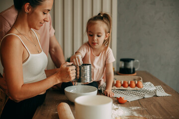 Happy family preparing dough together at home in the kitchen. A young brunette woman in a white top holds a flour sieve in her hands. A young brunette man in a pink T-shirt is watching the process.