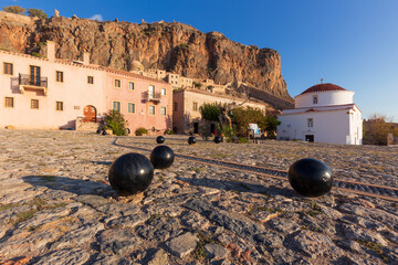 Cobbled square with white church and few cannonballs in the foreground at the foot of a high cliff