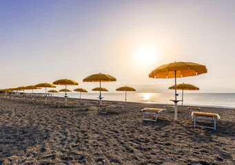 scenic view at nice beach with rows of umbrellas and chaise lounges with blue sea and amazing sunrise sky with sun glow on the background