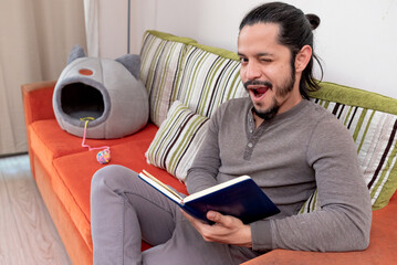 Young man reading a book at home sitting on the couch in the living room looking at camera