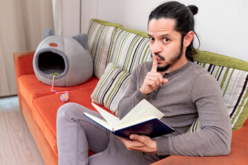 Young man reading a book at home sitting on the couch in the living room looking at camera