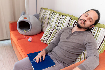 Young man sleeping while reading a book at home sitting on the couch in the living room