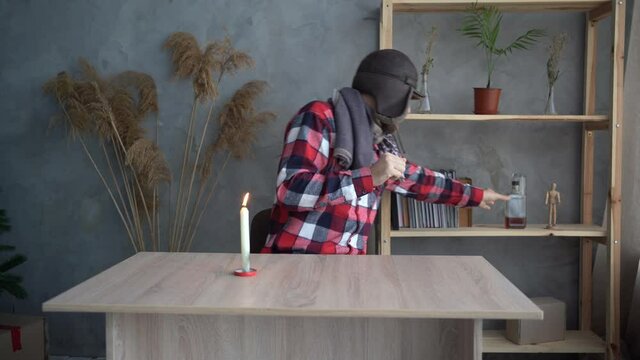 Bearded caucasian man in a hat at home warming himself with a candle and drinking whiskey, cold home concept