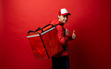 Delivery guy employee man in red cap and uniform workwear work as dealer courier hold red thermal food bag and showing thumb up isolated on red color background studio. Service concept.