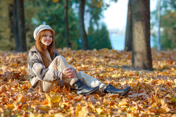 A young pretty girl with long red hair, lies in the fallen leaves in an autumn park among the trees, in a cap and jacket, and smiles. Blue sky and tall trees. Joy, autumn, relax.