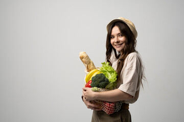Fototapeta premium Portrait of happy smiling young woman in beige oversize t-shirt holding reusable string bag with groceries over white background. Sustainability, eco living and people concept.