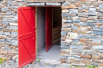 A historic cold storage vegetable cellar underground with two red wooden doors in the brick walls.  The building has a rock facing wall, concrete floor, and the roof is made of soil and sods.  