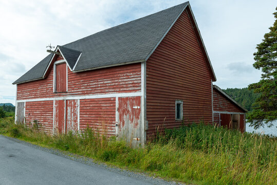 A vintage red two story barn with white trim, black shingled roof with multiple doors and windows. The old farm building is near water and has green grass surrounding the exterior of the wooden barn.