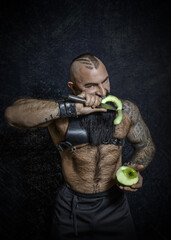 A brutal sexy man with a black beard with a naked hairy torso cuts a green apple with a large knife and eats a piece of apple