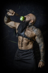 Brutal sexy man with a black beard with a naked hairy torso bites a green apple on a large knife