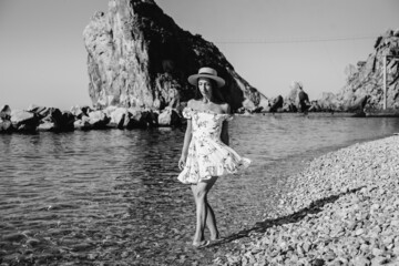 A beautiful young woman in a hat and a light dress is walking along the ocean shore against the background of huge rocks on a sunny day. Tourism and vacation travel. Black and white.