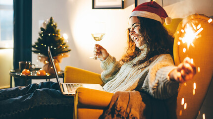 Happy young woman wearing Santa hat, drinking champagne, talking with friends on video call...