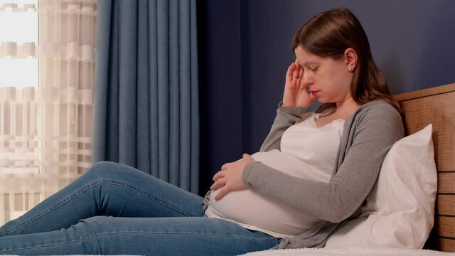 A pregnant woman in bed feels a headache and tries to calm her baby by stroking the belly. How to get rid of headaches when you are pregnant.