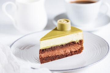 Pistachio chocolate cake with four layers and green pistachio glazing on top served on a white plate on white background with coffee in blur
