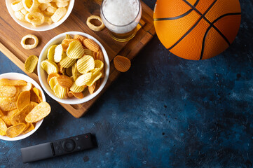 A classic set of sports fans - a glass of beer, potato chips, onion rings on a wooden tray. Nearby on the table is a television remote control and a basketball. watching sports matches on TV.