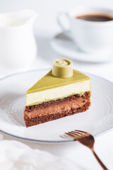 Pistachio chocolate cake with four layers and green pistachio glazing on top served on a white plate on white background with coffee in blur