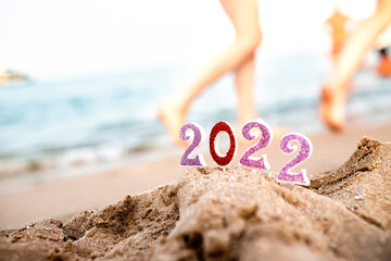 Figures 2022 on sand against background of sea beach at resort in southern countries with feet of vacationers walking on water at sunset and celebrating arrival of new year near ocean