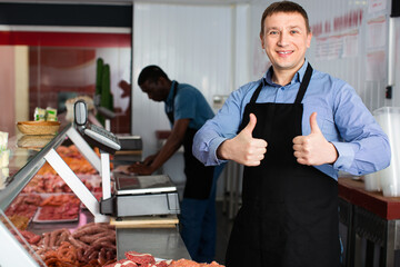 Proud cheerful positive smiling male owner of butcher shop standing behind counter, giving thumbs up