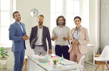 Team of creative business people during work meeting. Group portrait of happy young corporate employees standing by office desk with laptop computer and post it notes, smiling and looking at camera