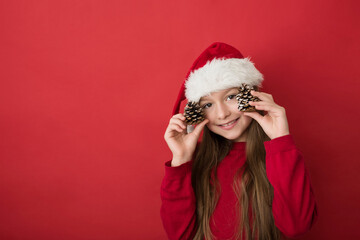 A little girl with long dark hair in a red Santa Claus hat smiles broadly, holding two fir cones in her hands.