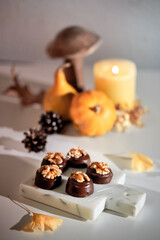 Chocolate pralines with walnut nuts on marble cutting board. Closeup on candy. Autumn arrangement with candle, pumpkins, wooden mushroom anddry yellow ginkgo leaves. Natural sunlight, with long