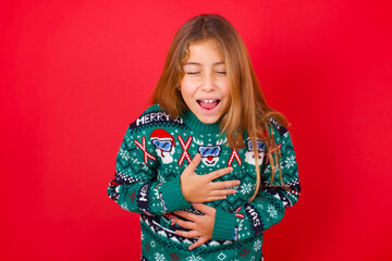 little kid girl wearing knitted sweater christmas over red background keeps hands crossed, laughs out loud at good joke, wears casual clothes.