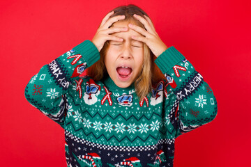 Shocked panic little kid girl wearing knitted sweater christmas over red background holding hands on head and screaming in despair and frustration.