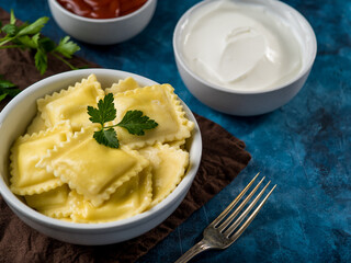 One serving of Italian ravioli with parsley leaf and two gravy - tomato and sour cream. There is a fork nearby. Dinner table for one person. Restaurant, hotel, cafe, home cooking.