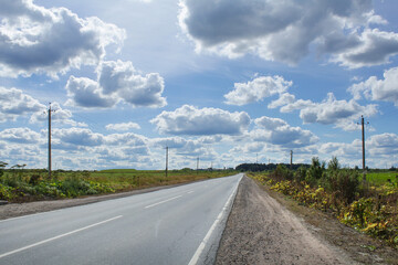 Fototapeta na wymiar Empty highway under blue sky with clouds on sunny day, poles with electric wires and green grass on the sides