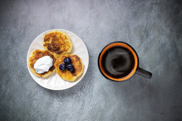 Three cheesecakes with blueberries and sour cream on white plate, black-orange cup of coffee with steam on gray table, view from above, flat lay