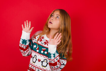 little kid girl wearing knitted sweater christmas over red background keeps palms forward and looks with fright above on ceiling tries to defense herself from invisible danger opens mouth.