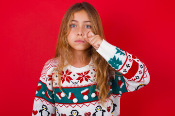 Disappointed dejected little kid girl wearing knitted sweater christmas over red background wipes tears stands stressed with gloomy expression. Negative emotion