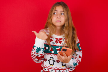 little kid girl wearing knitted sweater christmas over red background points thumb away and shows blank space aside, holds mobile phone for sending text messages.