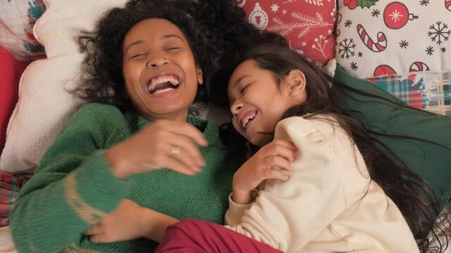 Black mother in knitted design sweater and daughter lie on pillows tickle kisses on nose hugging in Christmas atmosphere slow motion