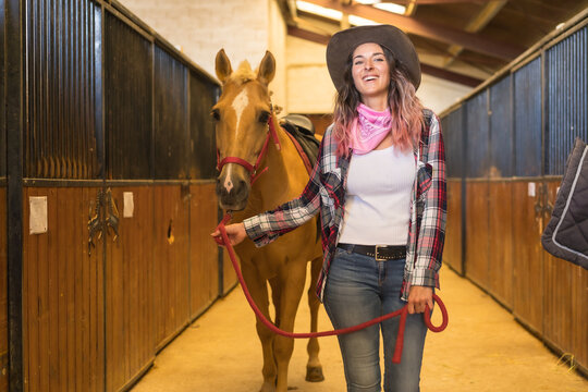Caucasian pink haired cowgirl woman strolling with a horse in a stable, American cowboy hats, pink plaid shirt and jeans