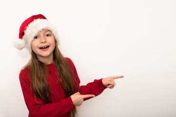 Little smiling girl in red Christmas sweater and red Santa hat points to a place for text or design with fingers isolated on white background, emotions on her face, wow