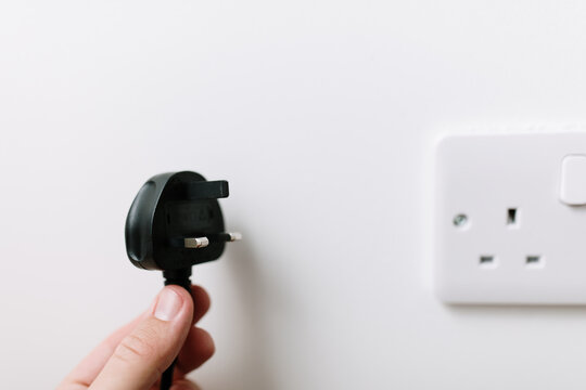 A hand is holding a black plug from the United Kingdom. Unplugged concept due to high energy prices. A tip for saving energy. A blurry socket in the background. White wall, natural light. Copy space.