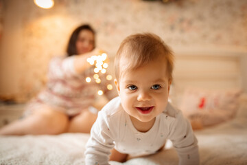 a child in white clothes is crawling on the bed and smiling. In the background, mom plays with her daughter and christmas lights