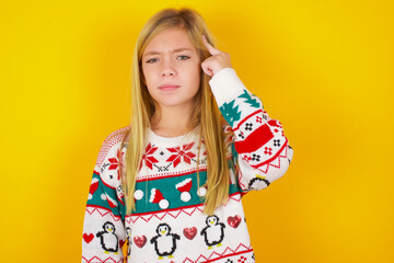 caucasian little kid girl wearing knitted sweater christmas over yellow background tries to memorize something, keeps fore finger on temple, reminds information for exam,