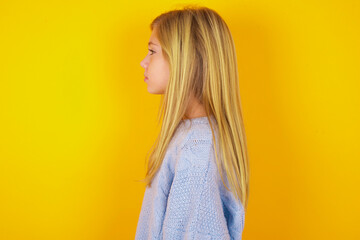 caucasian kid girl wearing blue knitted sweater over yellow background looking to side, relax...