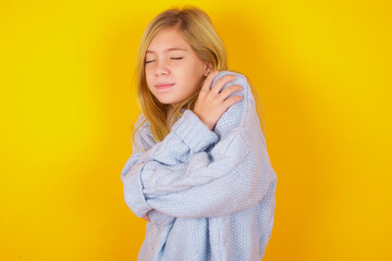 caucasian kid girl wearing blue knitted sweater over yellow background Hugging oneself happy and positive, smiling confident. Self love and self care