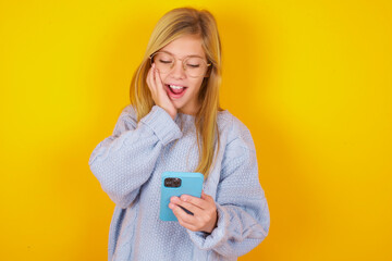 caucasian kid girl wearing blue knitted sweater over yellow background  holding in hands cell reading sms using new app 5g