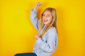 Profile photo of excited caucasian kid girl wearing blue knitted sweater over yellow background  raising fists celebrating black Friday shopping